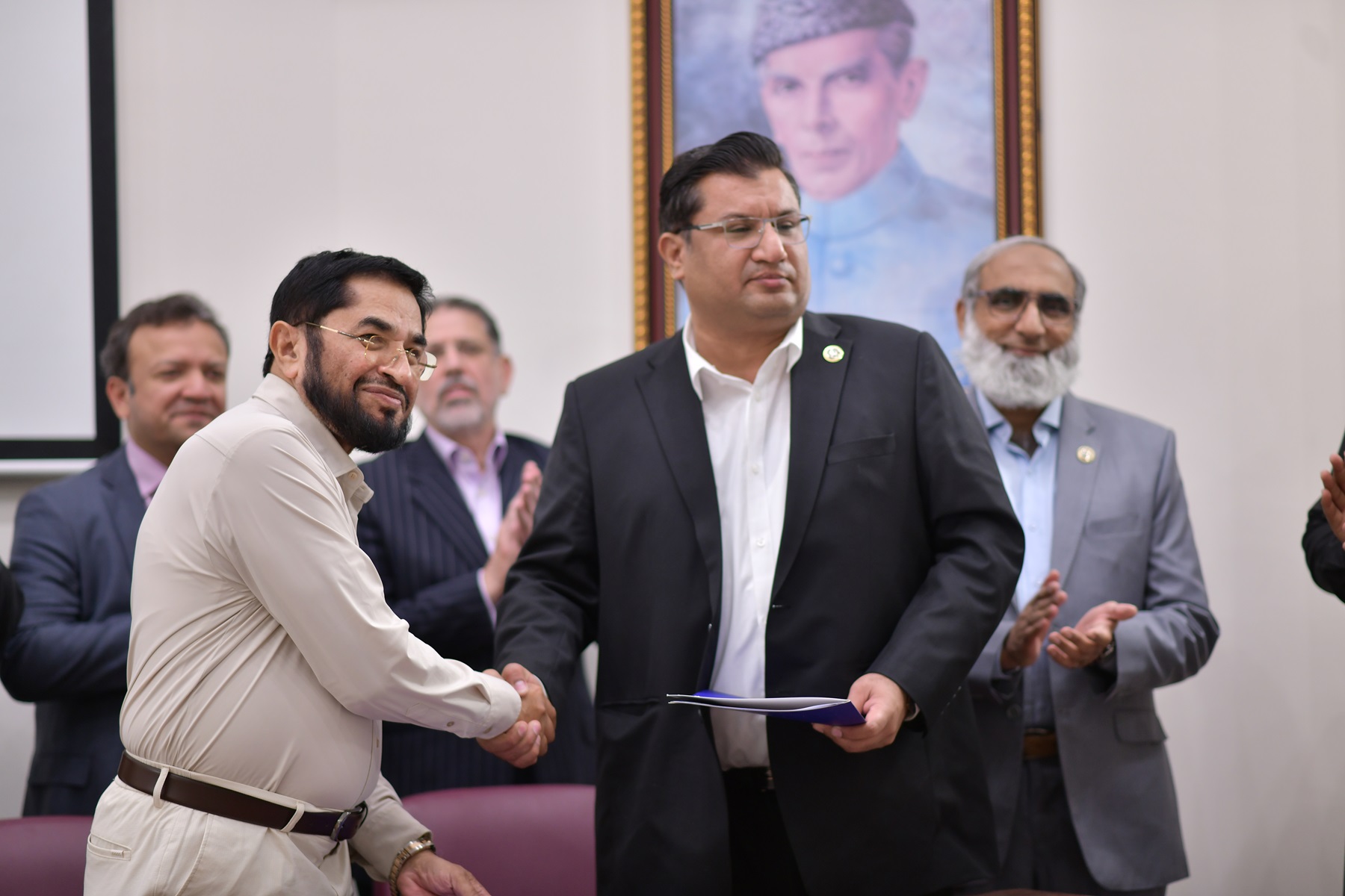 Certificate handover during formal ceremony; two men exchange document while others applaud in official setting.